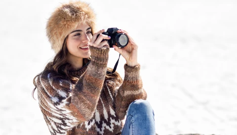 Woman in knit sweater and winter hat holding vintage camera to her eye while smiling.
