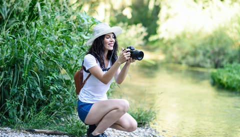 Woman crouching by a riverside holding a camera, wearing a white cap and casual clothing.