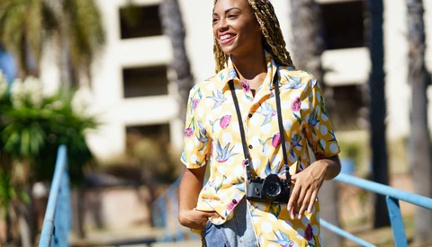 Smiling woman wearing a colorful floral shirt and camera on a lanyard, standing outdoors near palm trees.