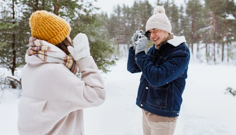 Two people playing in snow, one in white jacket and yellow hat, the other in denim jacket and white beanie, snowball fight in snowy forest.