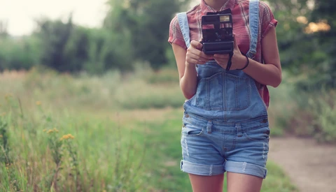 Person in denim overalls and plaid shirt holding a vintage camera in a rural field.