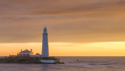 Lighthouse on rocky promontory at golden hour with keeper's house and outbuildings nearby.