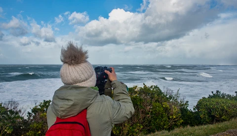 Person in winter hat and jacket photographing ocean waves from a coastal clifftop.