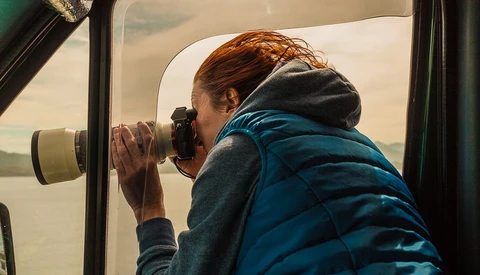 Photographer with reddish hair peers through binoculars from inside a vehicle window.