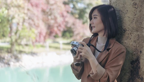 Woman holding a vintage rangefinder camera while leaning against a tree with blooming flowers in soft focus background.