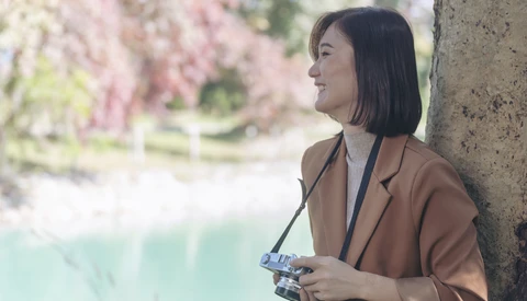 Woman holding a vintage camera, leaning against a textured wall with blooming flowers in soft focus behind her.