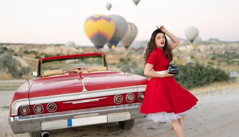 Woman in red dress posing beside vintage red convertible with hot air balloons rising in desert landscape.