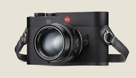 Leica rangefinder camera with textured black body and fixed lens, shown with attached shoulder straps against neutral background.