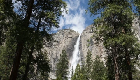 Waterfall cascading down granite cliffs framed by towering evergreen trees and blue sky.