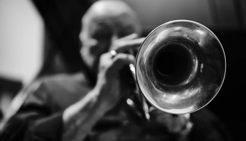 Musician playing trumpet, photographed from front with instrument bell in sharp focus and player blurred in background.