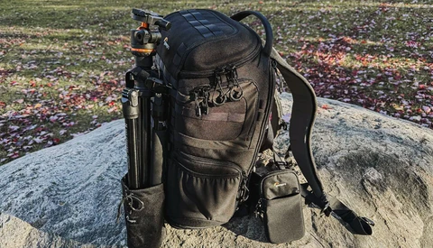 Black camera backpack with tripod attachment sitting on a rock in an outdoor field setting.