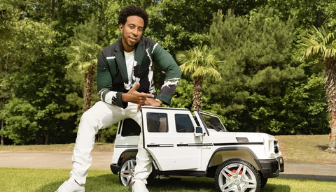 Man in green jacket and white pants posing with a miniature white SUV on grass with trees in background.