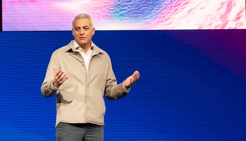 Man presenting on stage with hands gesturing while speaking before a blue backdrop with colorful digital art.