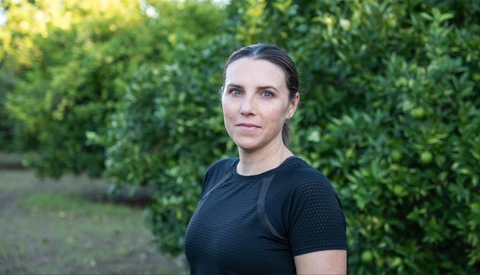 Woman in black t-shirt standing in front of green foliage, looking toward camera.