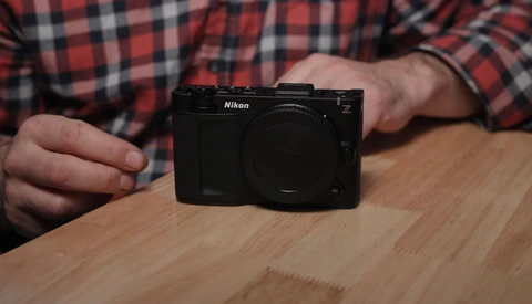 A black Nikon Z mirrorless camera body displayed on a wooden surface, held by a hand wearing a red plaid shirt.