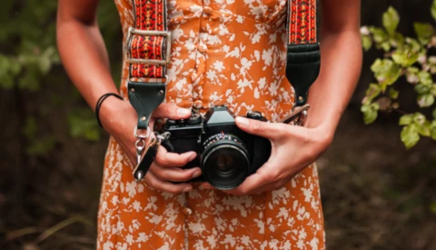 Photographer holding a vintage film camera with decorative shoulder strap, wearing an orange floral dress.