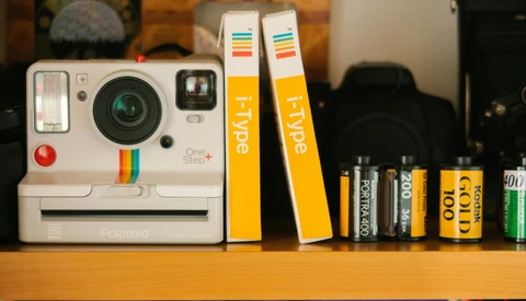 Polaroid One Step+ camera with rainbow stripe branding displayed on wooden shelf alongside Hype film boxes and various film cartridges.