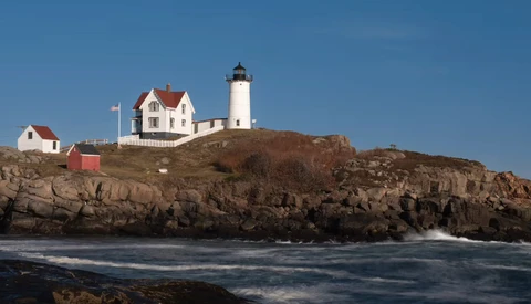 White lighthouse with keeper's house on rocky coastal headland under clear blue sky.