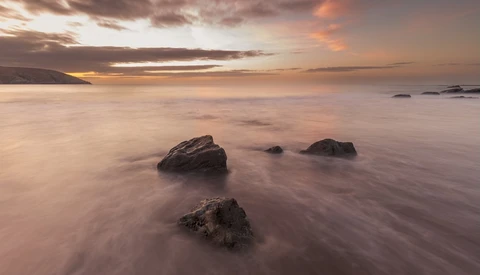 Rocky outcrops emerge from smooth water during golden-hour sunset with dramatic clouds overhead.