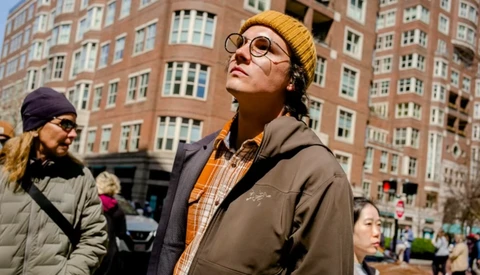 Person wearing glasses and yellow beanie looking upward on a city street with brick buildings behind.