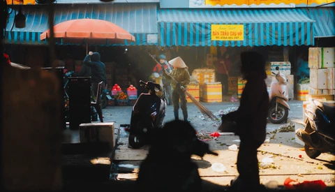 Street market scene viewed through silhouetted foreground figures, with vendors and blue corrugated metal structures under warm daylight.