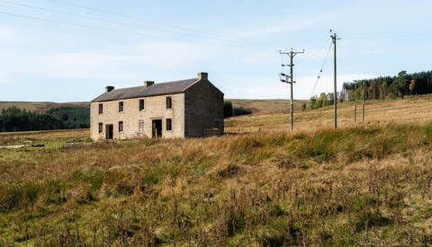 Abandoned stone farmhouse in overgrown moorland with utility poles under overcast sky.