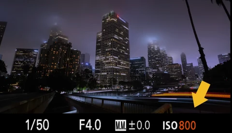 Urban skyline at dusk with illuminated skyscrapers reflected in still water, with camera settings displayed as an overlay.