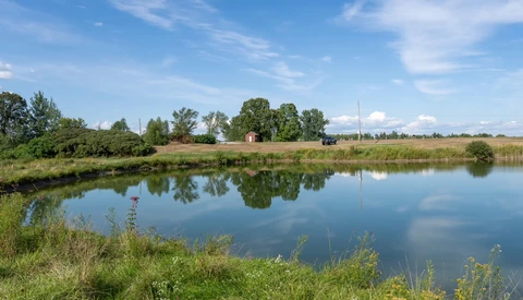 Rural landscape with calm river reflecting blue sky, trees, and distant buildings along grassy banks.