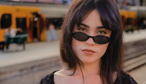 Woman wearing dark oval sunglasses and dark clothing at a train station platform.