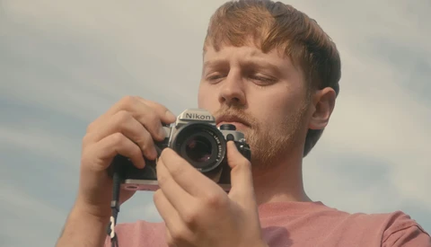 Male photographer holding a Nikon camera to his face while looking downward against a cloudy sky.