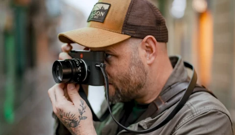 Photographer holding a rangefinder camera up to his eye while wearing a tan and brown trucker cap.