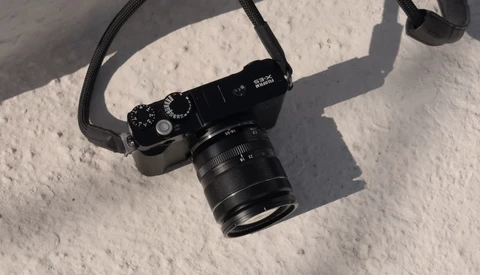Black rangefinder camera with attached lens on white textured surface, casting sharp shadows in bright sunlight.