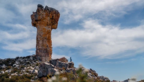 Tall rock tower formation rising from a rocky hillside with a small human figure standing nearby for scale.