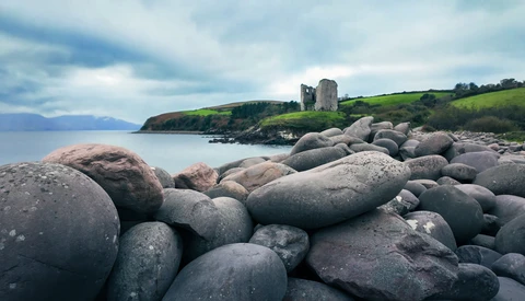 Boulder-strewn beach with castle ruins on distant headland under dramatic cloudy sky.