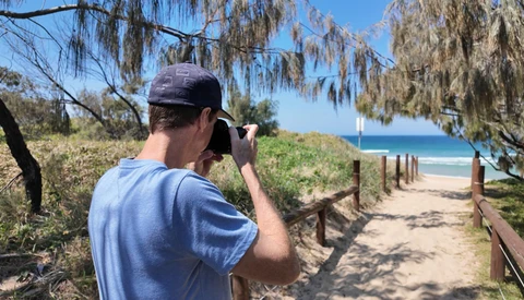 Photographer with camera raised to face, composing a shot down a sandy beach path lined with wooden railings and coastal vegetation.