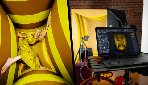 Photographer posing in yellow fabric setup while editor views the same shot on laptop screen nearby.