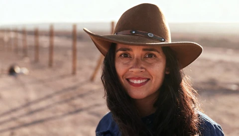 Woman wearing a brown cowboy hat and blue shirt, smiling at the camera in a desert landscape at golden hour.