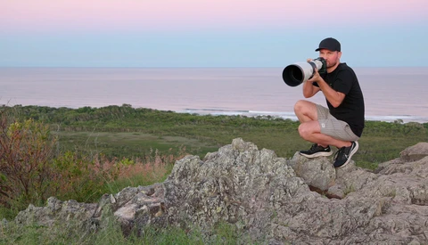 Photographer crouching on rocky outcrop while shooting with telephoto lens toward distant shoreline.
