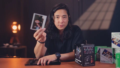 Creator holding instant film photograph at desk surrounded by camera equipment and lighting.