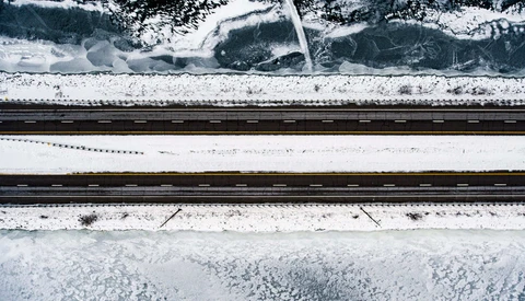 Aerial view of snow-covered landscape with railroad tracks and forested areas separated by white fields.