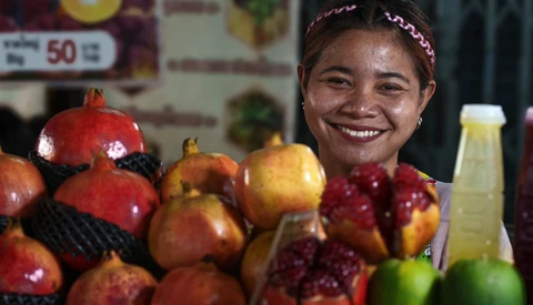 Smiling vendor at a fruit market stand displaying pomegranates and other fresh produce.