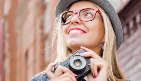Young woman wearing pink-framed glasses and gray beanie holding vintage camera, smiling upward against brick wall backdrop.