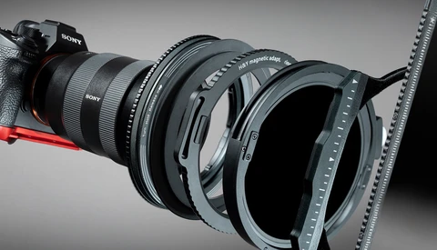 Sony mirrorless camera with multiple lens filters and adapters arranged in front of it against a gray background.