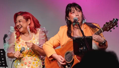 Two performers on stage with microphones; one in a yellow polka dot dress with red hair, the other in an orange jacket playing an acoustic guitar.