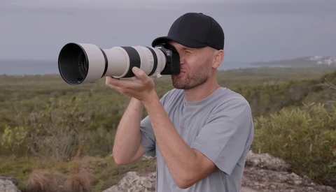 Photographer using a telephoto lens in an open landscape with scrub vegetation.