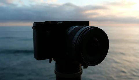 Compact camera with telephoto lens mounted on tripod, silhouetted against ocean and sunset sky.