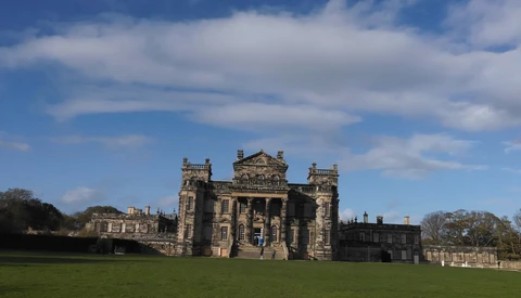 Stone castle with crenellated towers set on manicured grounds under partly cloudy sky.