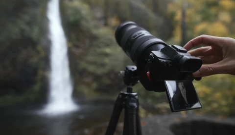 Photographer holding a mirrorless camera on a tripod with a waterfall in the background.