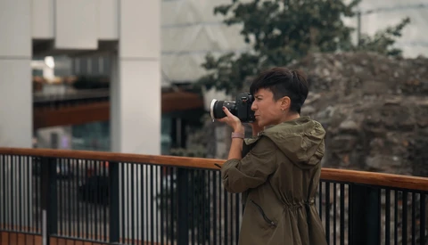 Photographer in olive jacket holding camera to eye while standing at railing overlooking urban architecture.
