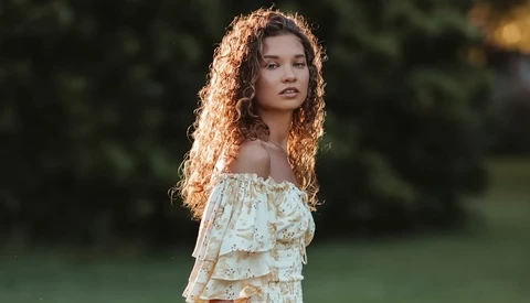 Young woman with long curly copper-toned hair wearing an off-shoulder ruffle blouse in a garden setting.